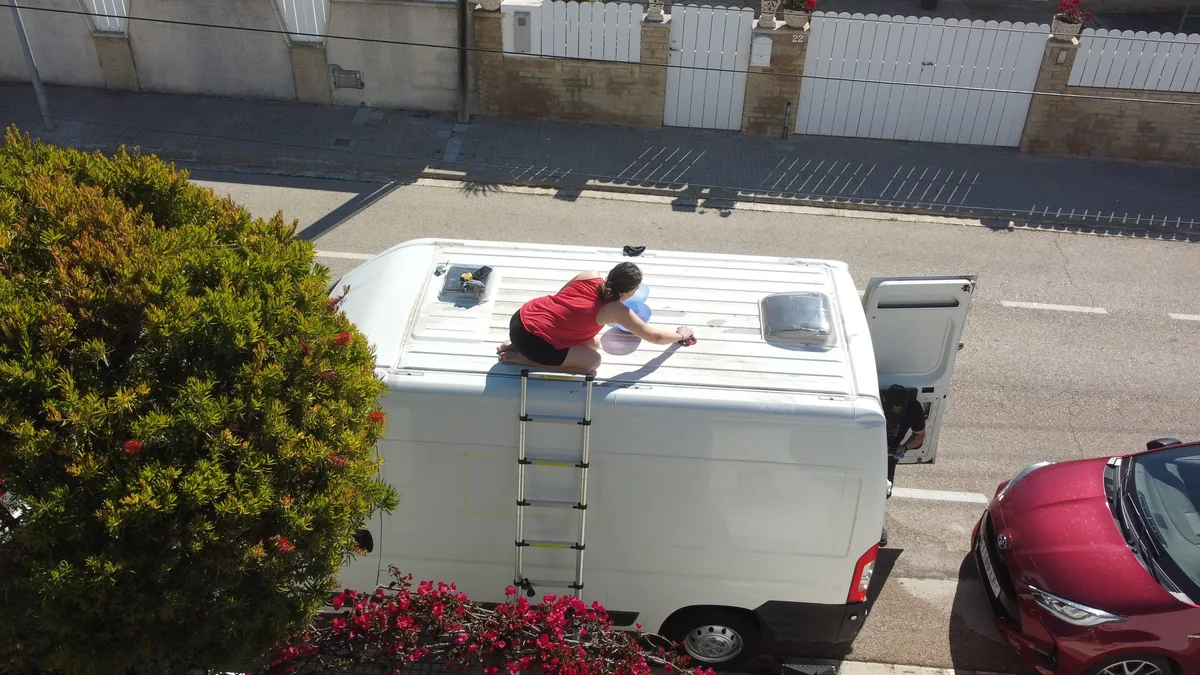 Preparación overland: mujer sobre furgoneta camper blanca limpiando techo y claraboyas con escalera en la calle