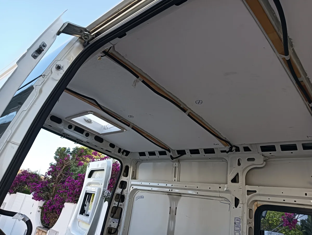 Interior de furgoneta camper en construcción: techo con aislamiento y claraboya, cableado eléctrico visible y flores moradas