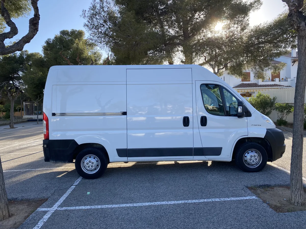 Furgoneta Peugeot Jumper blanca aparcada bajo pinos en un día soleado, perfecta para transformar en camper overland