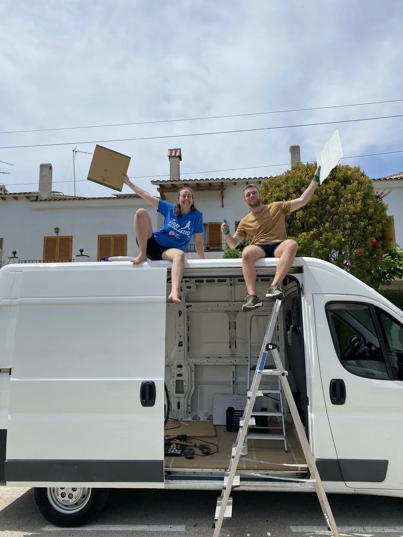 Pareja feliz sobre furgoneta blanca en plena camperización para viaje overland, con escalera y herramientas a la vista