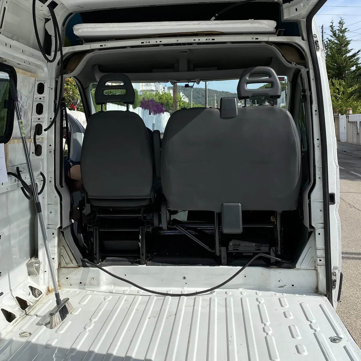 Interior de furgoneta blanca con asientos traseros, escoba y suelo metálico. Zona de carga preparada para camperizar