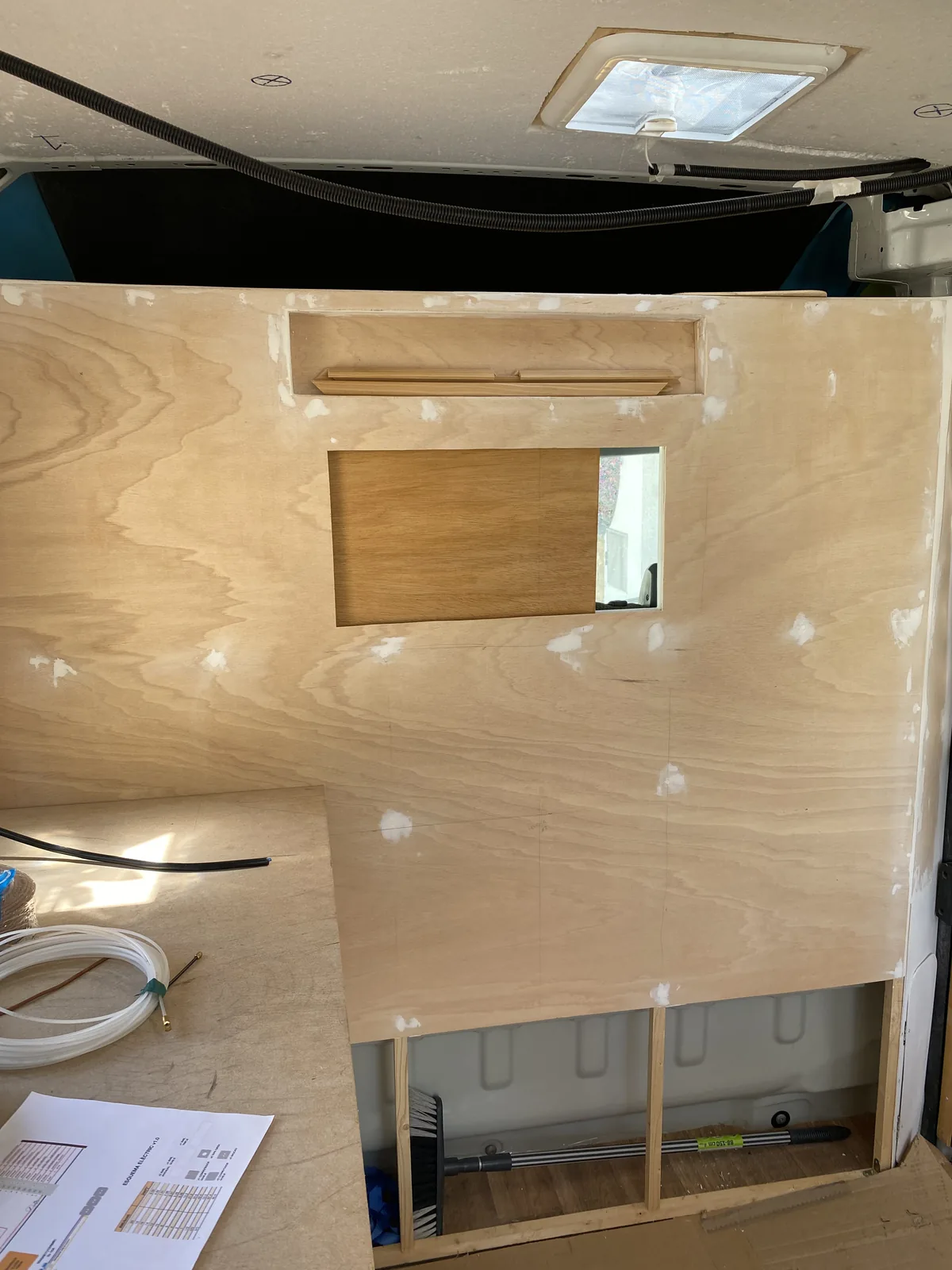 Interior de furgoneta camper en construcción, mostrando paneles de madera contrachapada, claraboya, cableado y esquemas