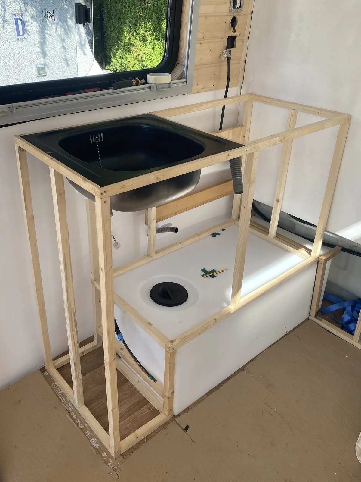 Cocina en construcción dentro de una furgoneta camper, con fregadero negro, depósito de agua y estructura de mader