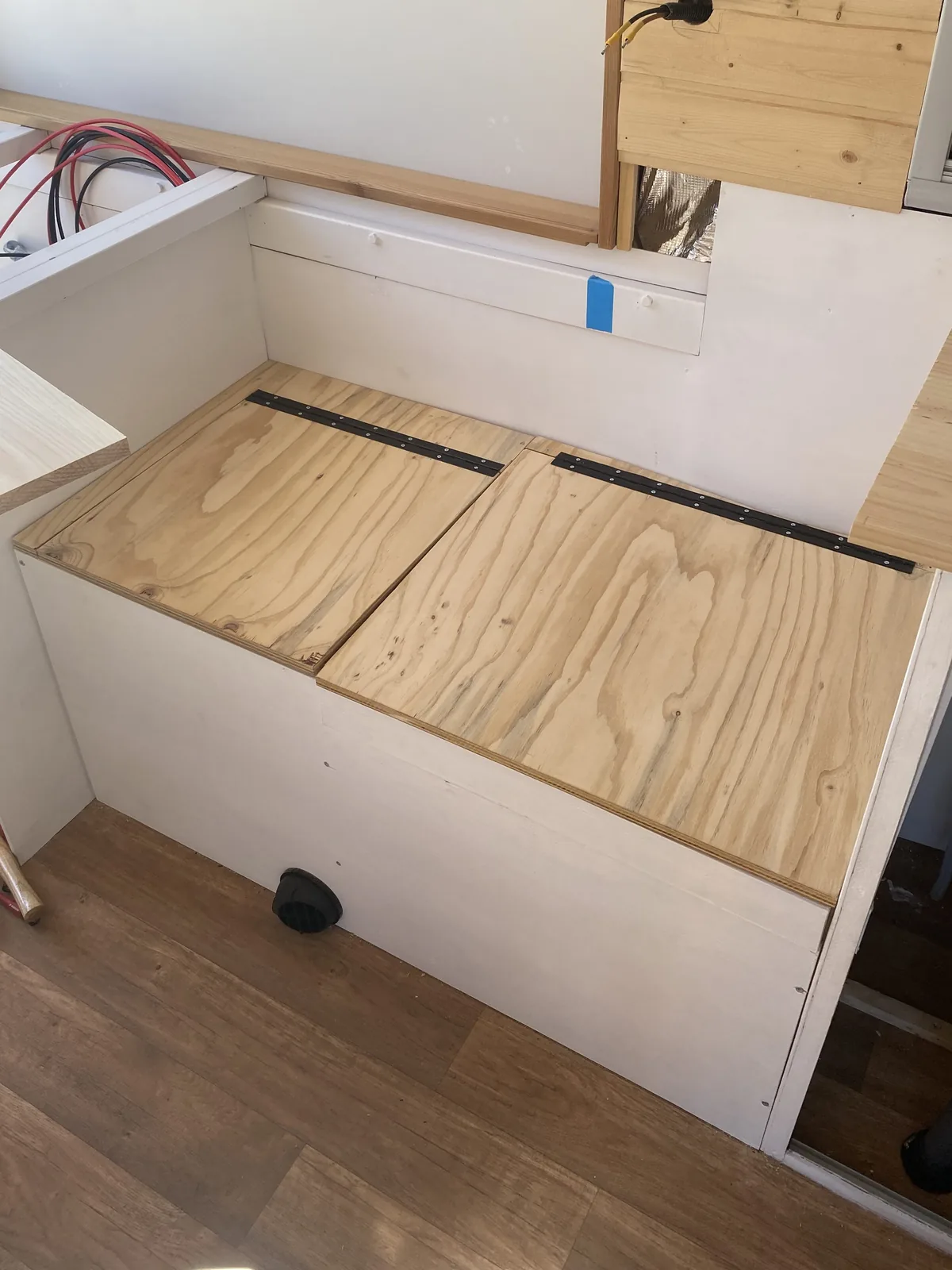 Interior de furgoneta camper en construcción con banco de almacenaje de madera y acceso a instalación eléctrica