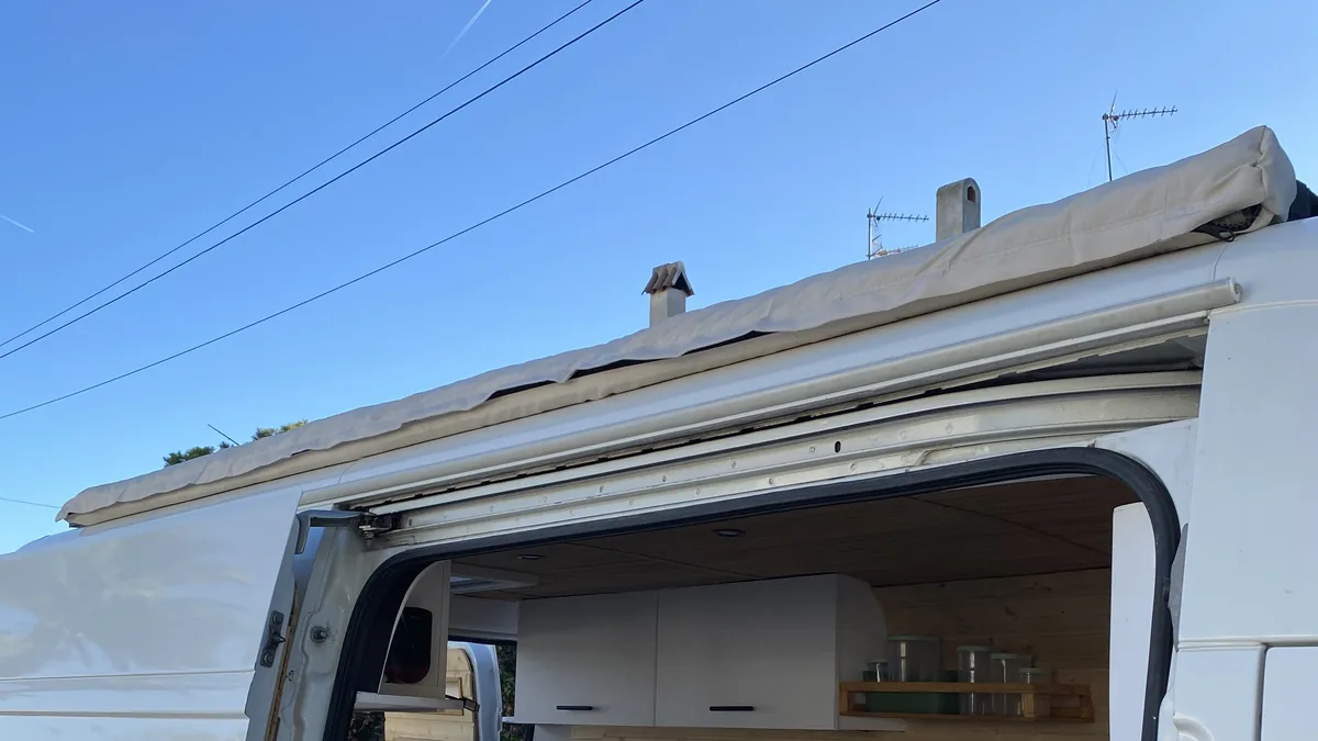 Furgoneta camper blanca con toldo enrollado, chimenea y antenas en el techo. Puerta lateral abierta a cocina interior
