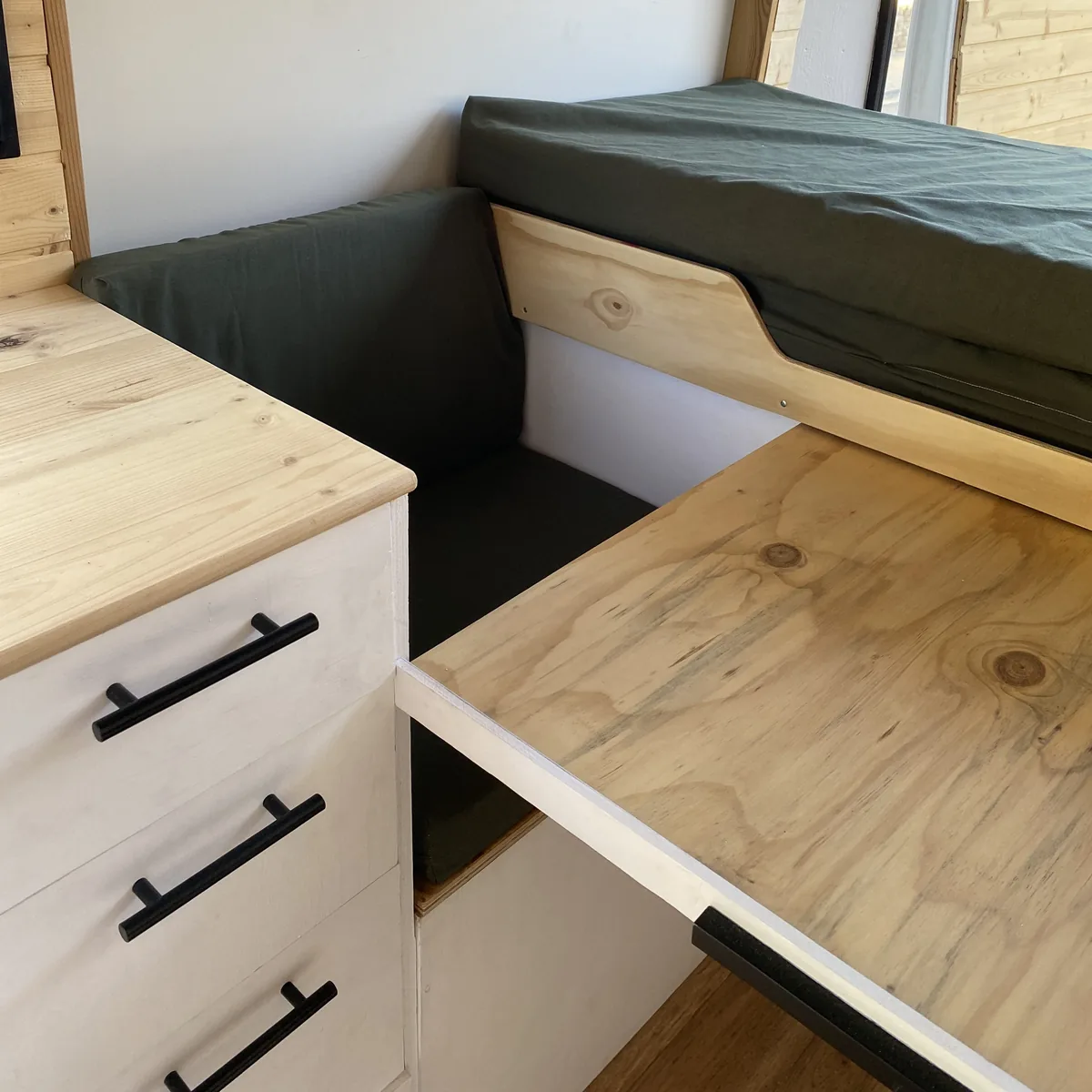 Furgoneta camper overland con interior de madera: cama elevada, asiento con cojines verdes y mesa, más cajonera blanca