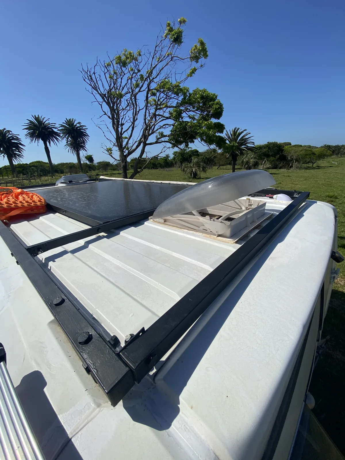 Techo de camper van overland con panel solar negro y claraboya abierta, en un paisaje verde con palmeras y cielo azul