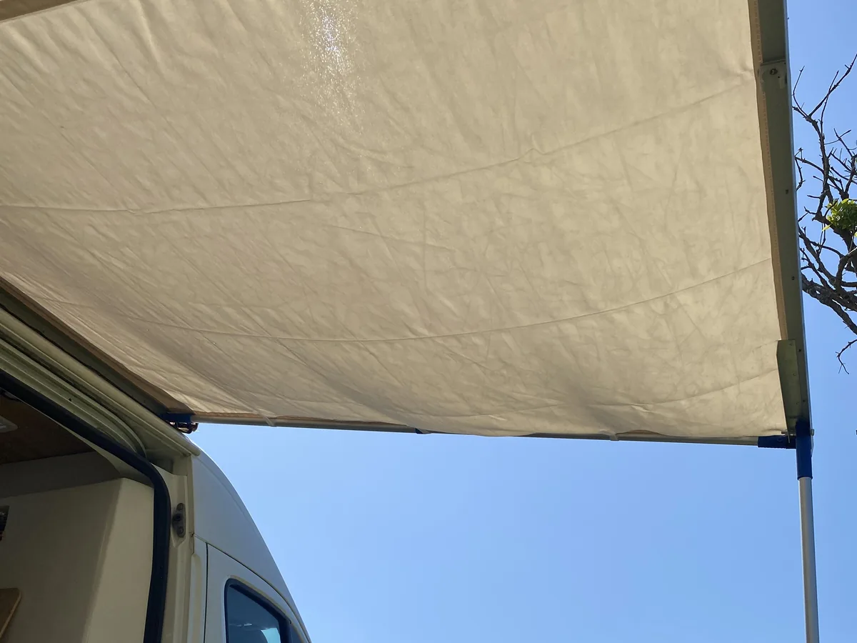 Toldo beige desplegado de una furgoneta camper, ofreciendo sombra bajo un cielo azul en un día de viaje overland