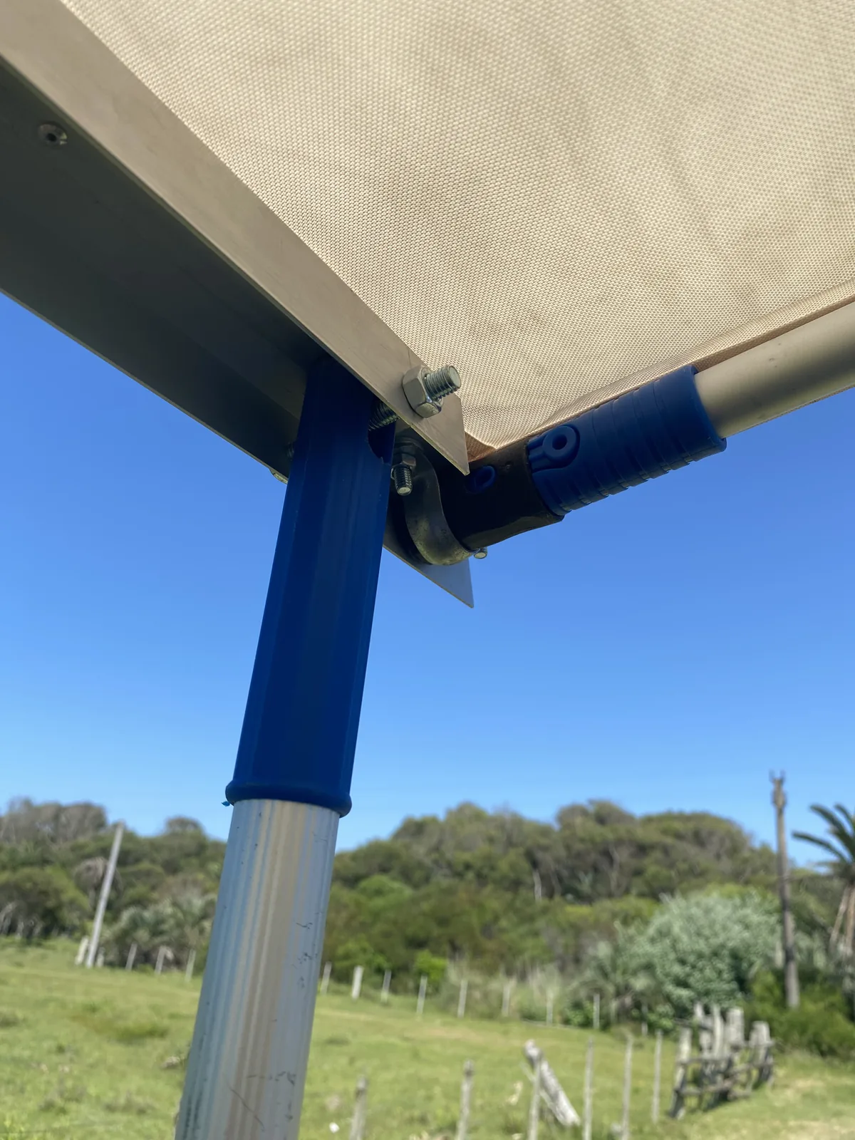 Detalle de anclaje de toldo de camper van con poste azul, en día soleado de viaje overland, campo verde y árboles al fondo
