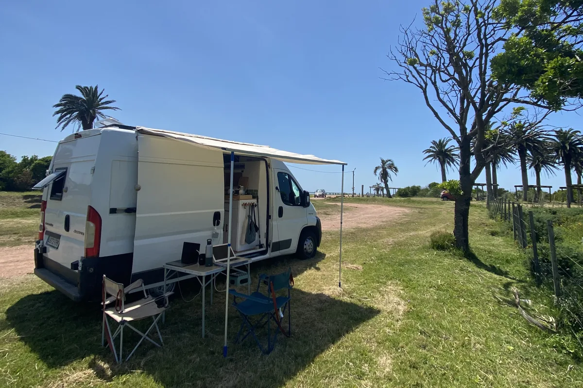 Furgoneta camper blanca con toldo, mesa y laptops afuera en un día soleado, rodeada de palmeras y cerca de la costa