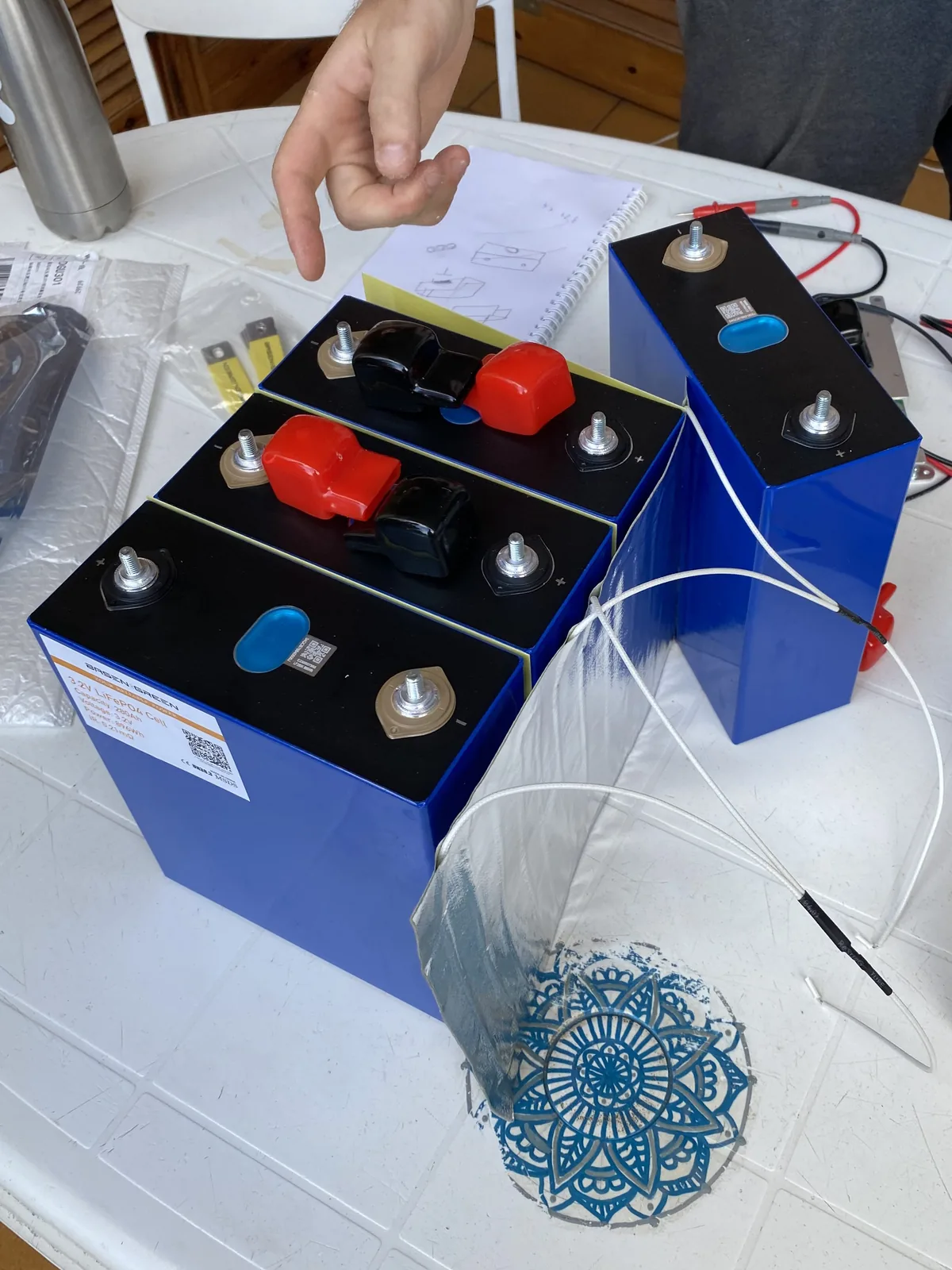 Preparación de celdas LiFePO4 azules con bornes protegidos para sistema eléctrico DIY en van camper, mano señalando