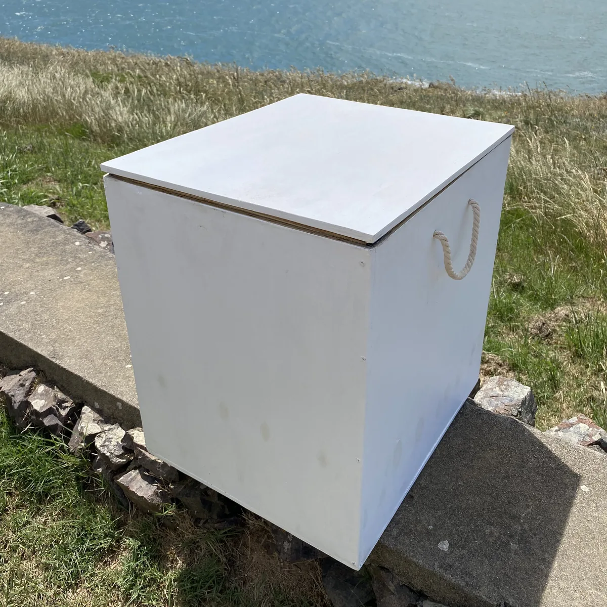 Caja de madera blanca con asa de cuerda sobre un muro, con el mar y vegetación costera de fondo