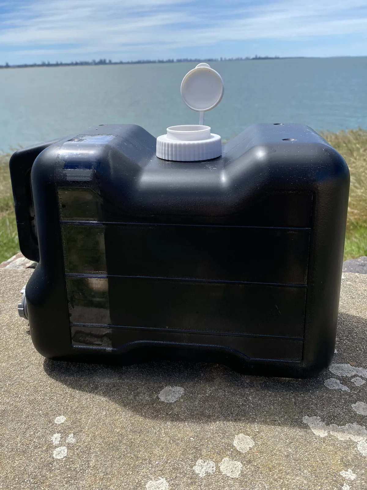 Bidón de agua negro con tapa blanca abierta para overland, sobre una roca frente al mar y una ciudad en el horizonte