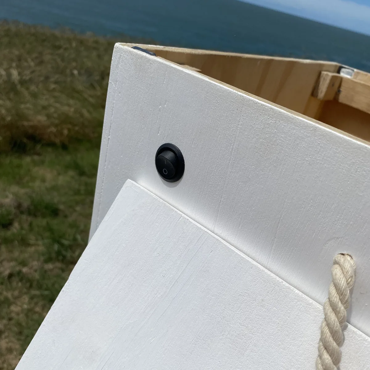 Interruptor negro en mueble de madera blanca para camperización, con el mar y vegetación costera de fondo