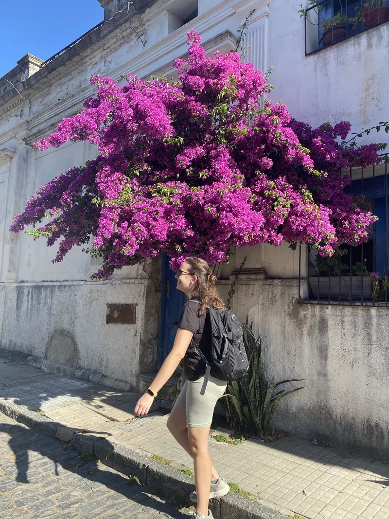 Viajera con mochila explorando una pintoresca calle colonial con adoquines, casas blancas y vibrantes buganvillas moradas