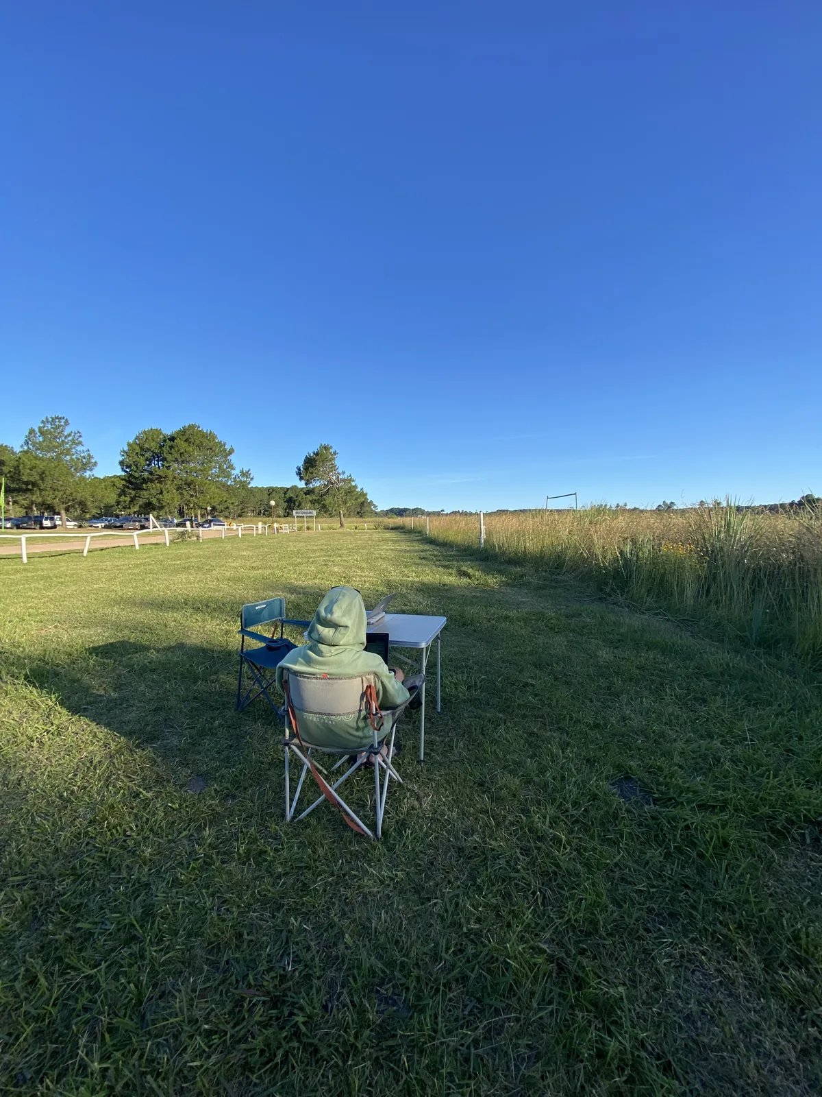 Viajero overland trabajando en su laptop desde una mesa de camping en un extenso campo de pasto bajo un cielo azul