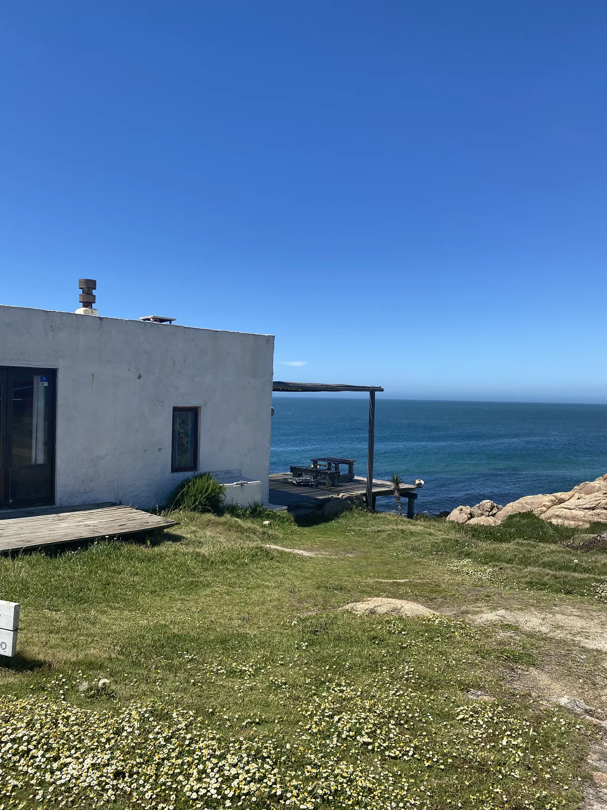 Cabaña rústica costera con deck de madera sobre el mar, rodeada de pasto florido y rocas bajo cielo azul