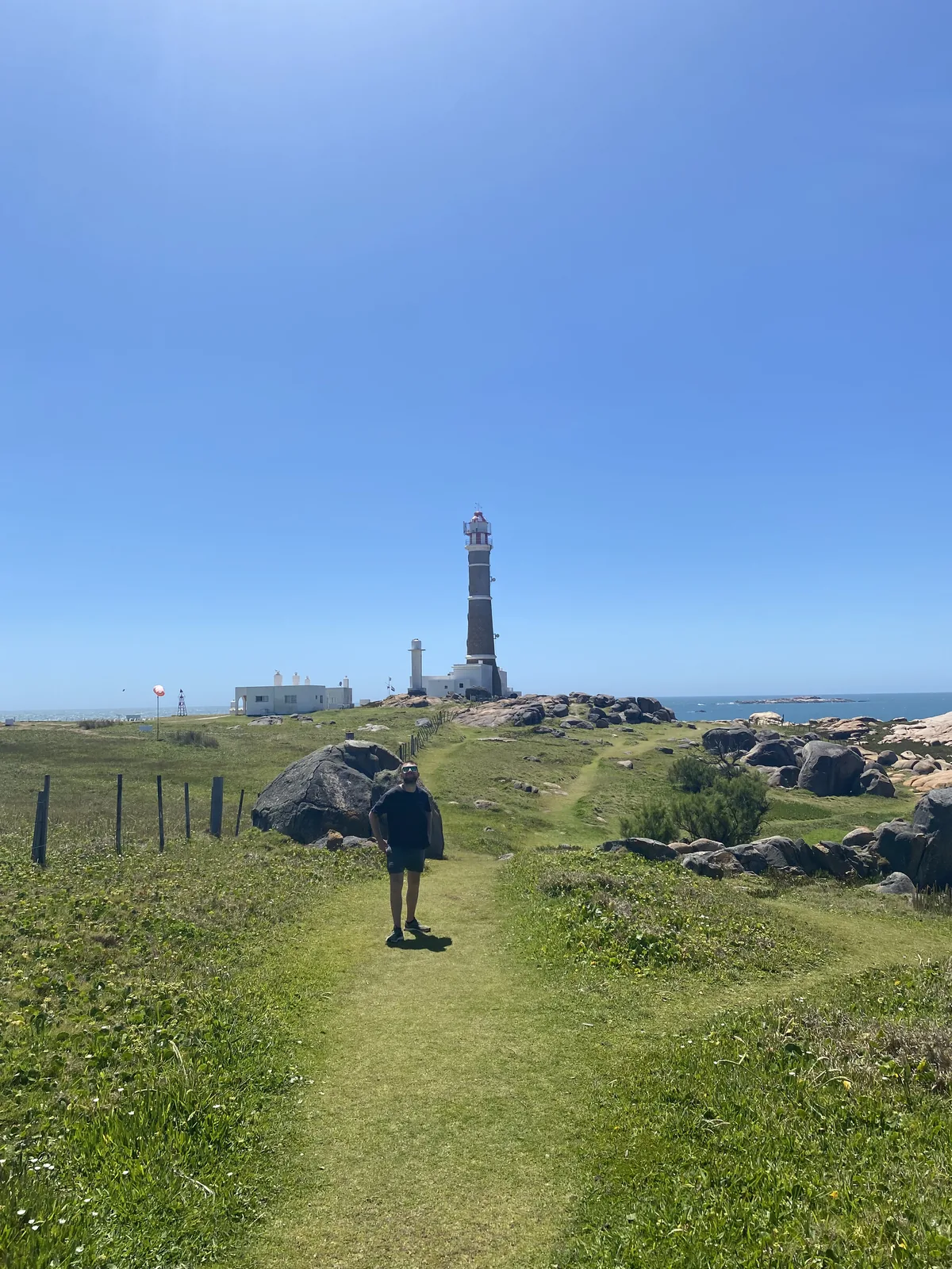 Hombre en sendero de pasto, explorando la costa rocosa de Cabo Polonio con su emblemático faro bajo sol