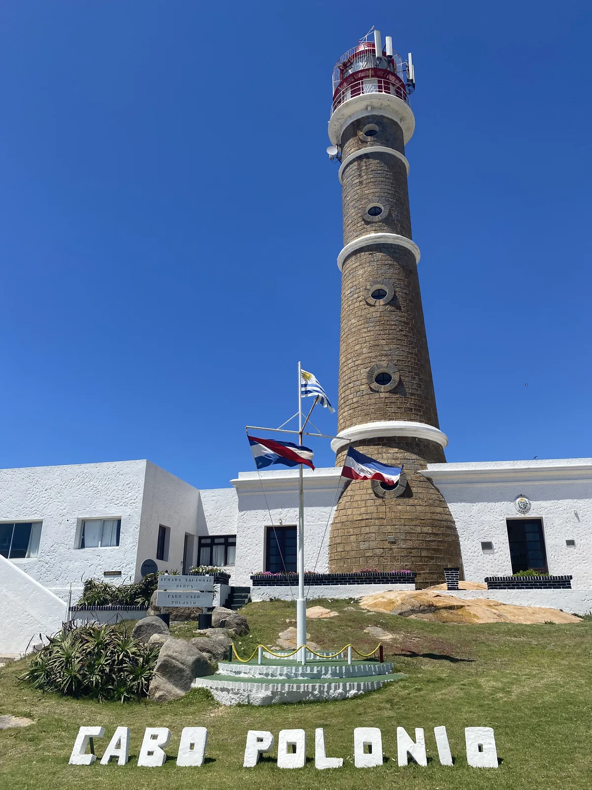 Día soleado en Cabo Polonio, Uruguay. El histórico faro de piedra se alza junto a edificios blancos y el cartel CABO POLONIO