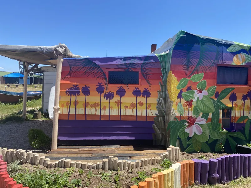 Caseta colorida con mural de palmeras al atardecer y plantas tropicales en un destino costero, con valla de troncos arcoíris