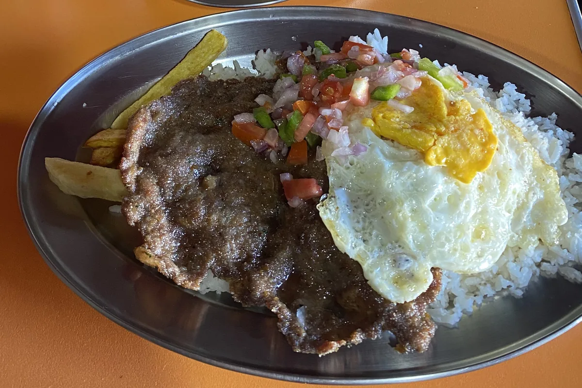 Comida local en plato de metal: bistec apanado, arroz, huevo frito, papas fritas y salsa criolla durante viaje overland