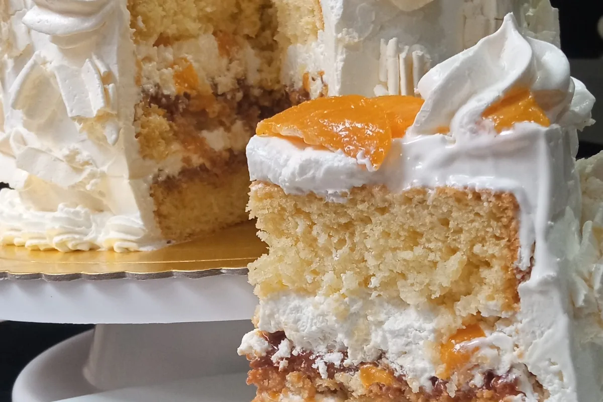 Pastel de capas con bizcocho claro, crema batida, un dulce relleno intermedio y trozos de durazno en la parte superior