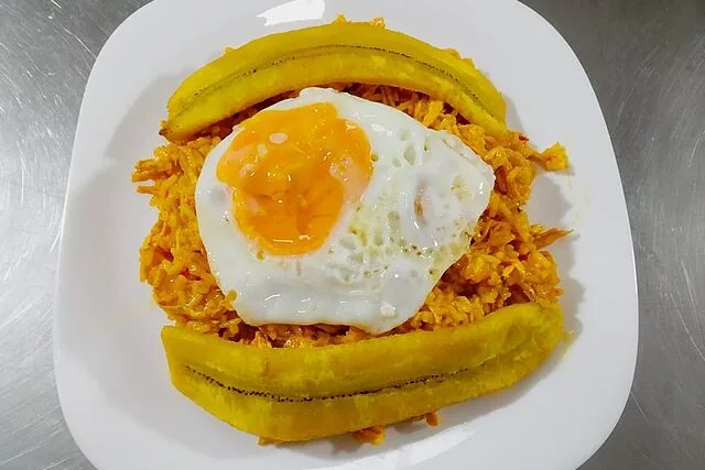 Típico plato de viaje: arroz amarillo, huevo frito y dos tajadas de plátano maduro sobre mesa metálica