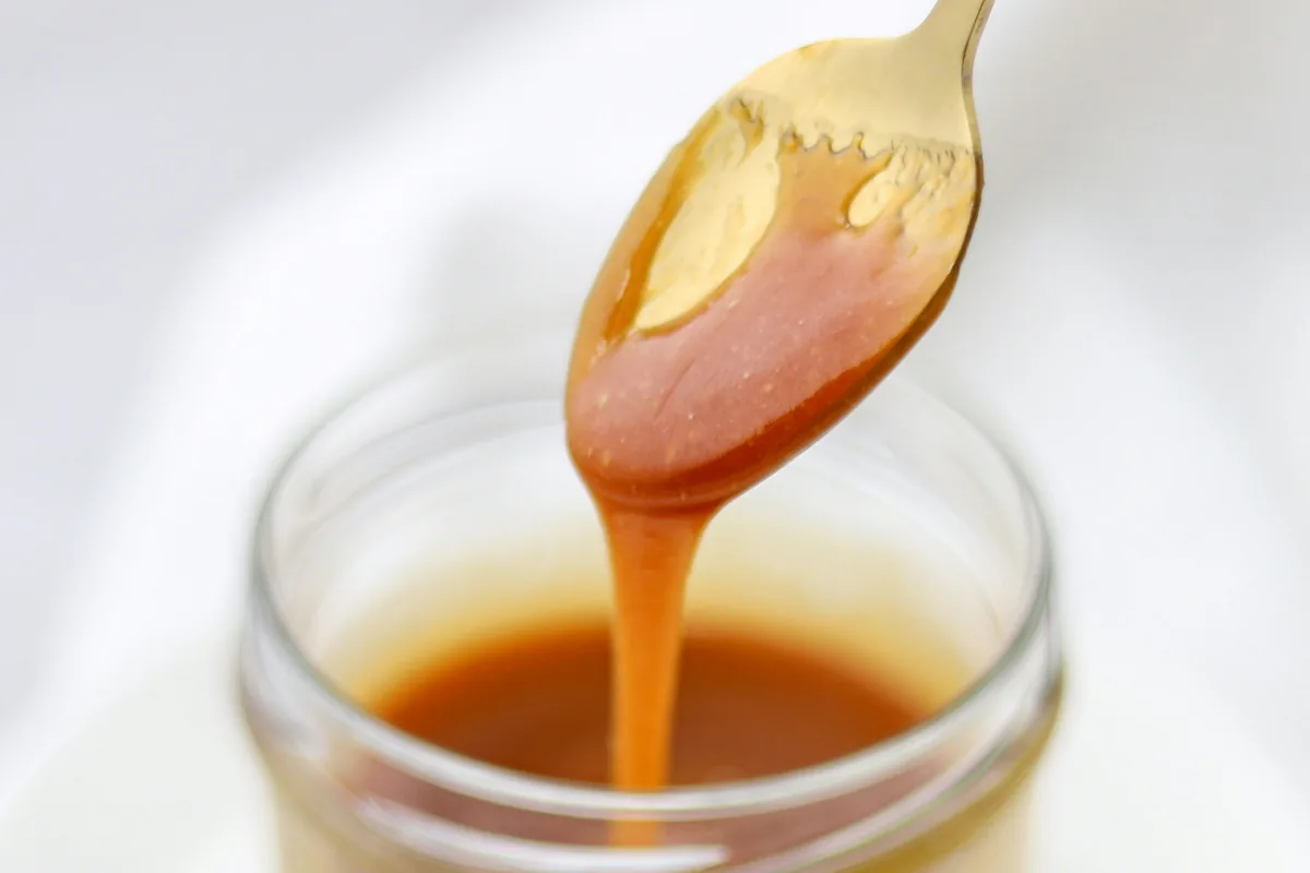 Cucharada dorada con caramelo espeso goteando en un frasco de vidrio, con un fondo blanco borroso