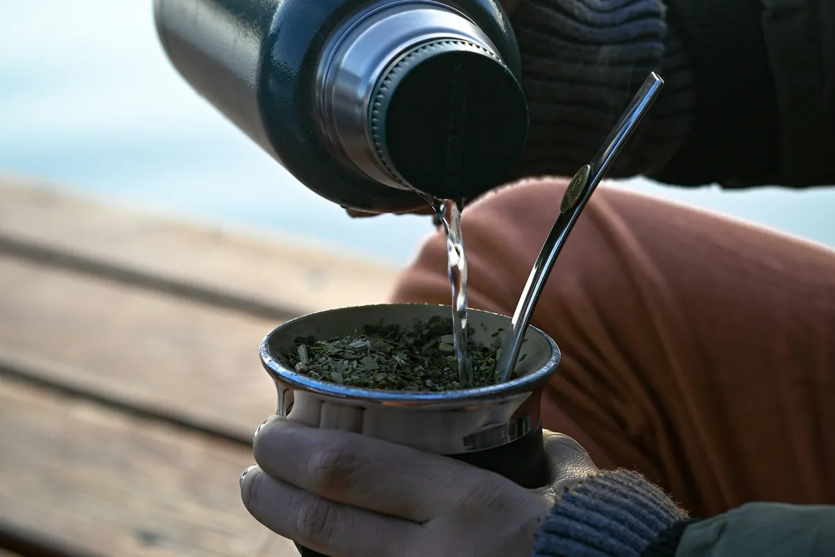 Manos de viajero preparando mate con termo Stanley verde sobre una tabla de madera, reflejo de agua