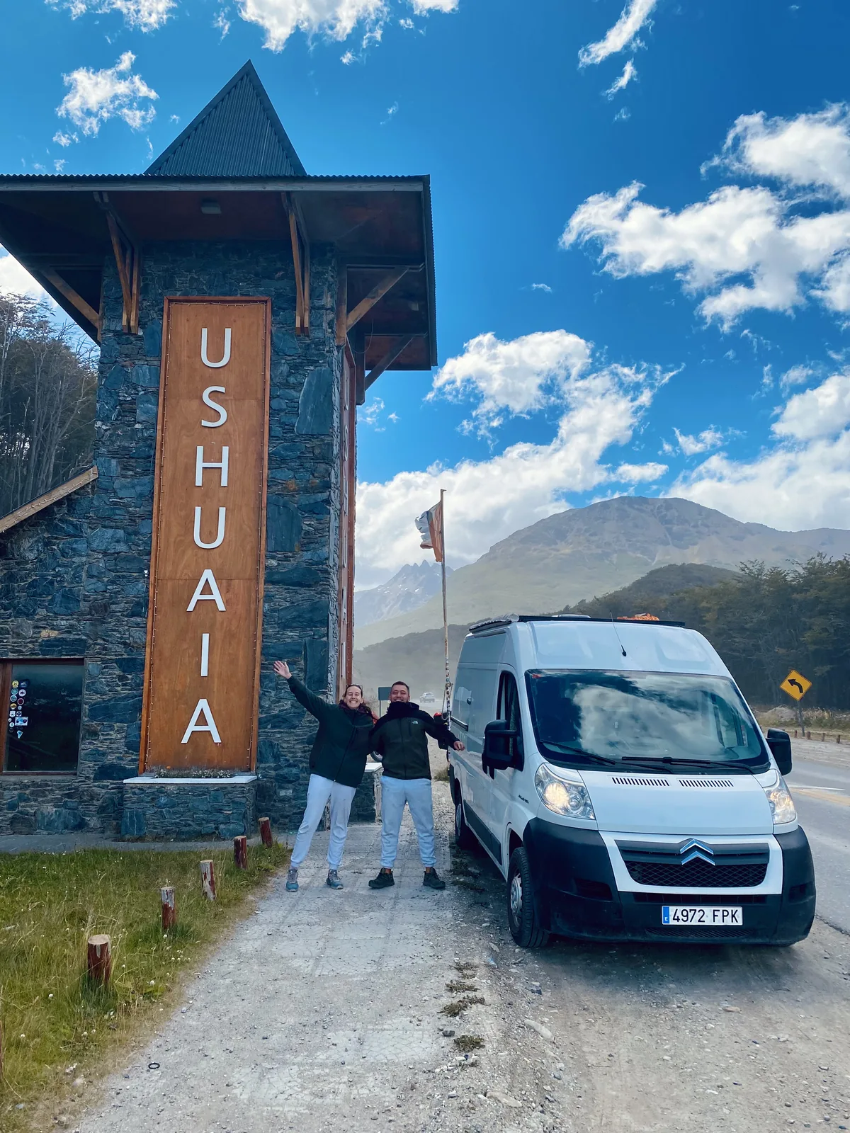 Dos overlanders celebran su llegada a Ushuaia posando con su van camper frente al cartel y montañas patagónicas