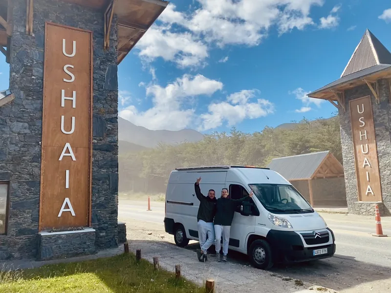 Pareja celebra con su furgoneta camper Citroën en el portal de bienvenida a Ushuaia, Fin del Mundo, montañas patagónicas