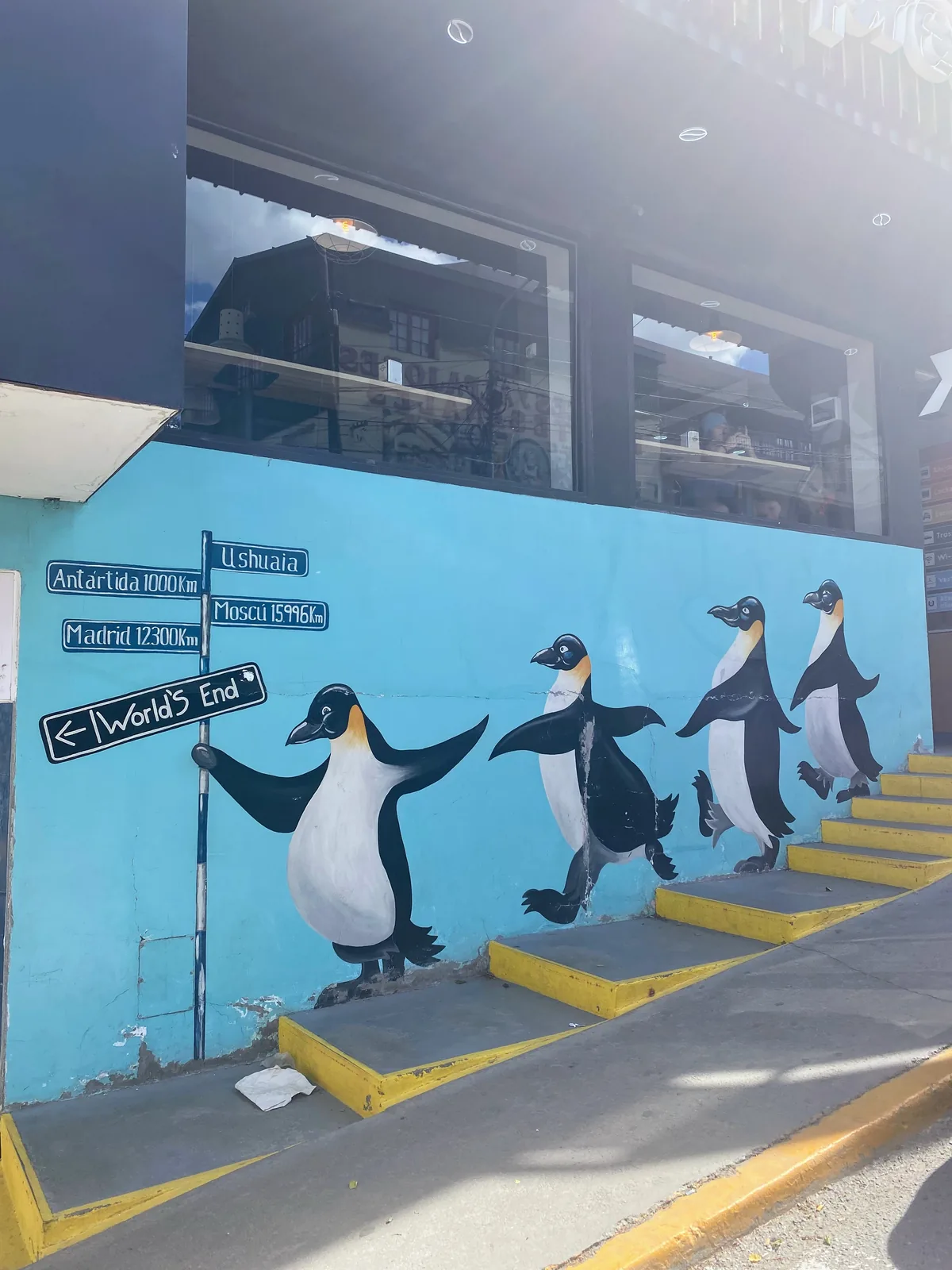 Mural de pingüinos en Ushuaia con carteles indicando la Antártida, Madrid y el Fin del Mundo, junto a escalones amarillos
