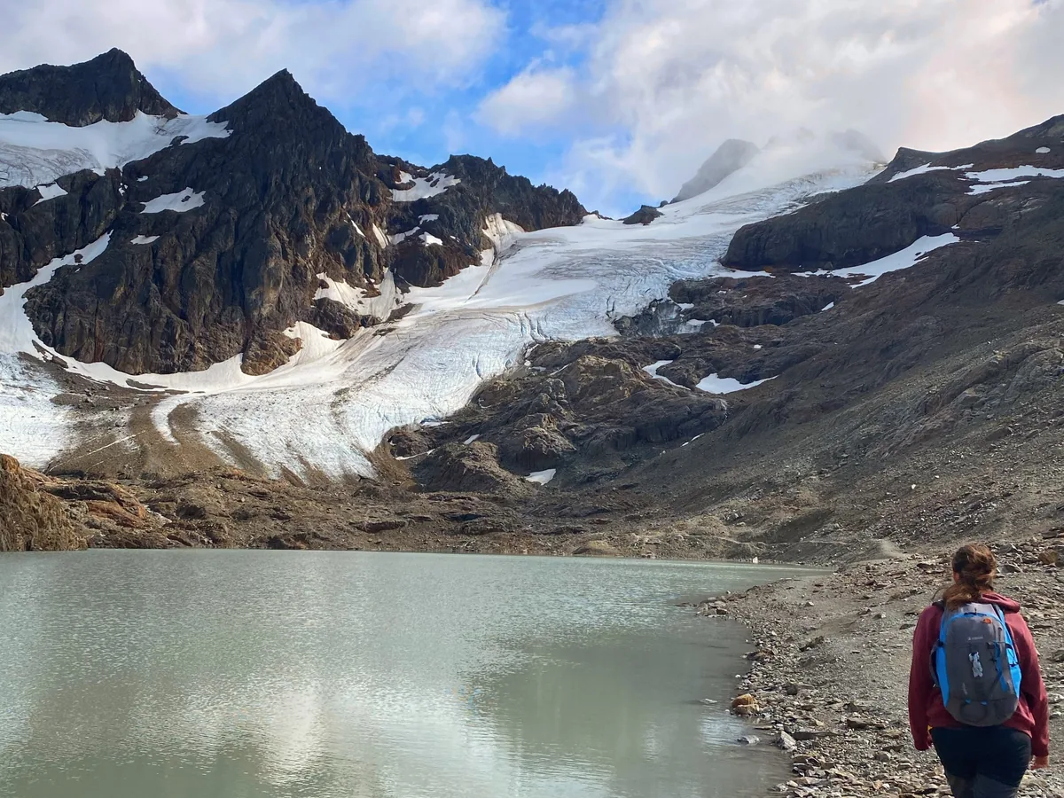 Mujer con mochila senderismo en la orilla de un lago glaciar con montañas nevadas y glaciares al fondo