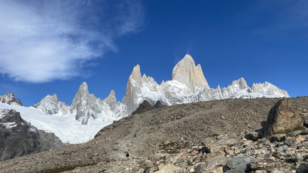 Imponente Fitz Roy y macizo de Cerro Torre en Patagonia, Argentina. Senderistas en primer plano rocoso bajo cielo azul.