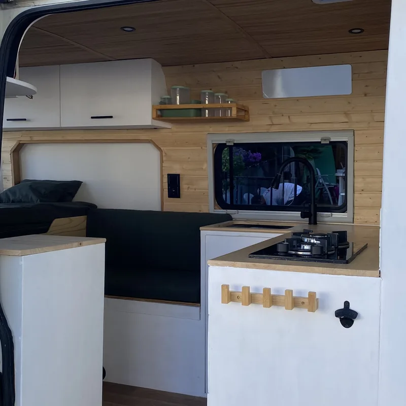 Moderna camperización de furgoneta overland: cocina con fogones y fregadero, cama y banco con cojines verdes, todo en madera