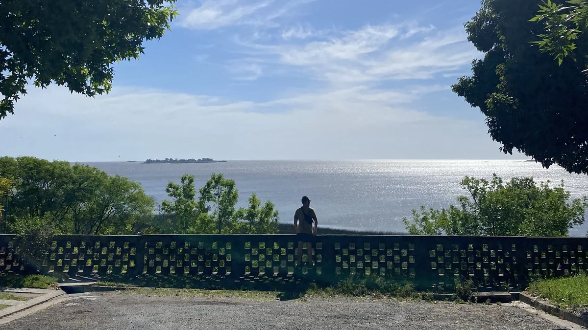 Viajero en un mirador con muro de diseño, observando un inmenso río o estuario brillante y las islas del horizonte.