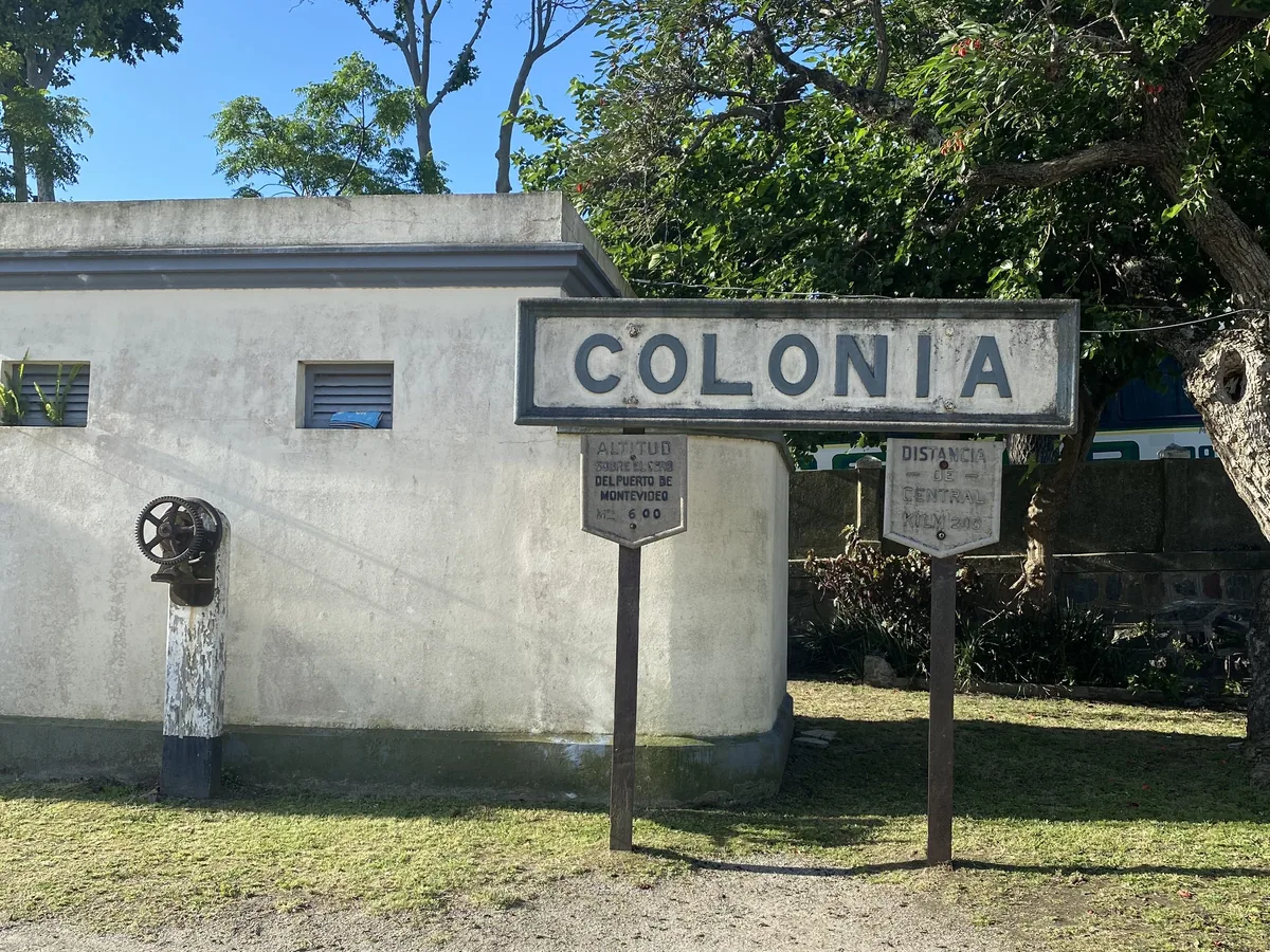 Panel de bienvenida a Colonia, Uruguay, con señales de altitud y distancia al puerto de Montevideo, entre árboles