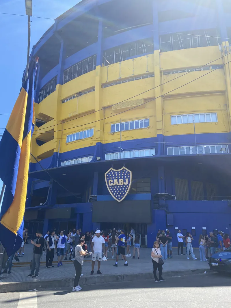 Fachada azul y amarilla del estadio La Bombonera en Buenos Aires, con bandera de Boca Juniors y gente en la entrada