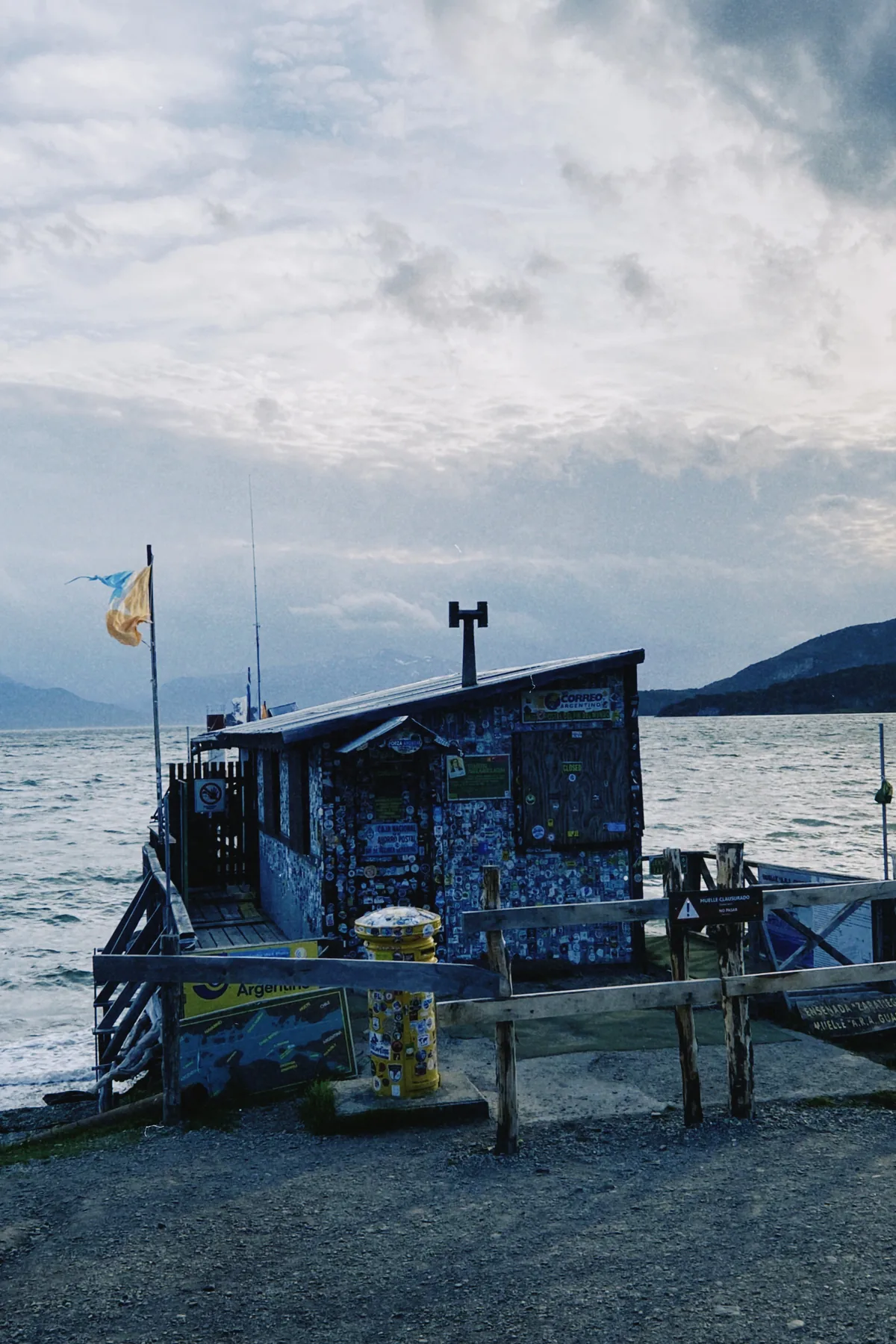 Casita de la Oficina Postal del Fin del Mundo en Bahía Ensenada, Tierra del Fuego. Buzón amarillo, mar agitado y montañas