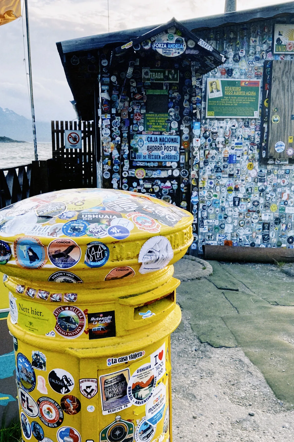 Buzón amarillo y cabaña llenos de stickers de viajeros overland en Ushuaia, Argentina, con montañas y el Canal Beagle