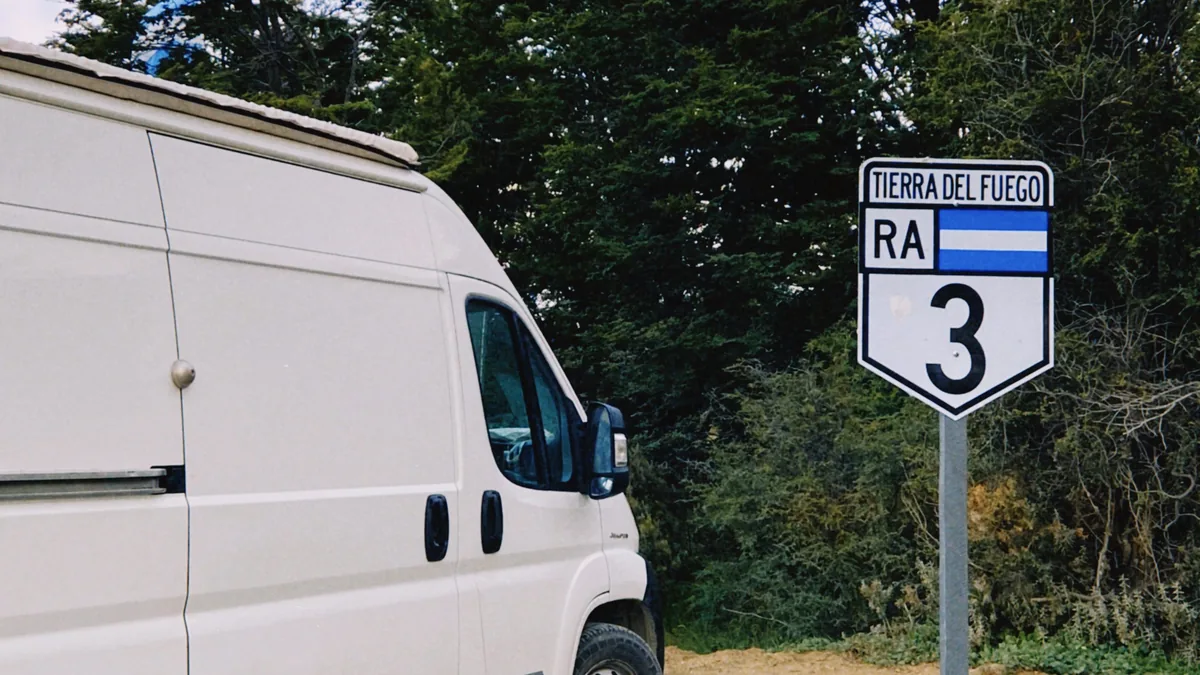 Furgoneta camper blanca estacionada junto a la icónica señal de Ruta 3 en Tierra del Fuego, Argentina, rodeada de bosque
