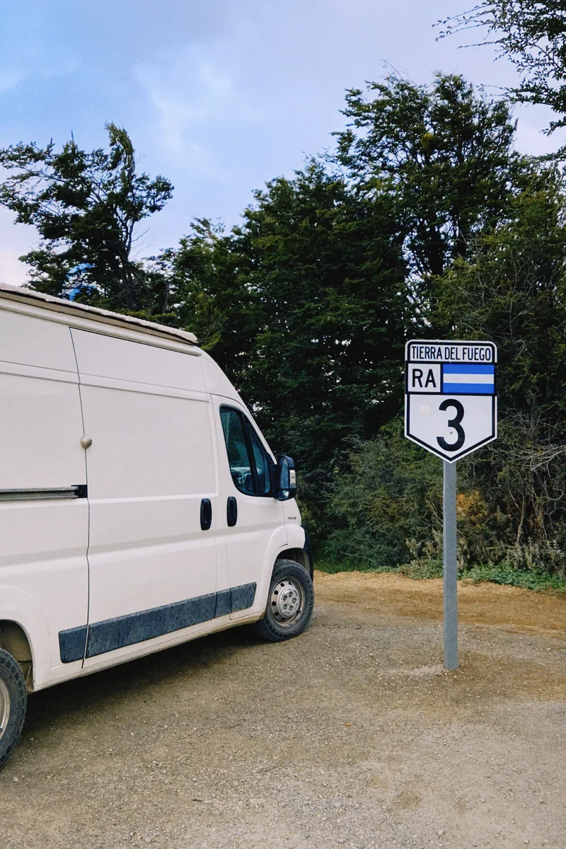 Furgoneta camper blanca junto a la señal de la Ruta Nacional 3 en Tierra del Fuego, rodeada de árboles