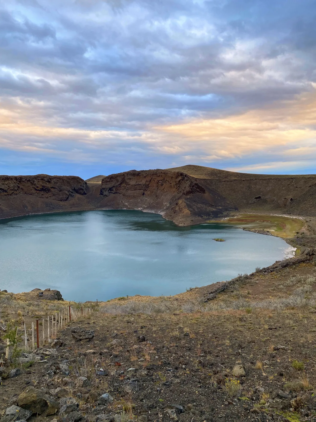 Laguna de cráter azul-verdosa rodeada de terreno volcánico árido, bajo un cielo nublado con tonos cálidos del atardecer