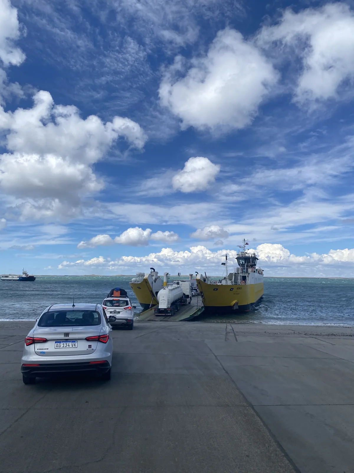 Coches de overland en rampa, incluyendo un Fiat Cronos y SUV con equipaje, esperando abordar dos ferries amarillos en el mar