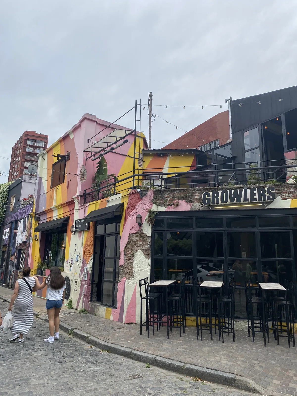 Barrio con fachadas coloridas y calle adoquinada. El bar Growlers con mesas en la acera. Dos mujeres pasean un día nublado