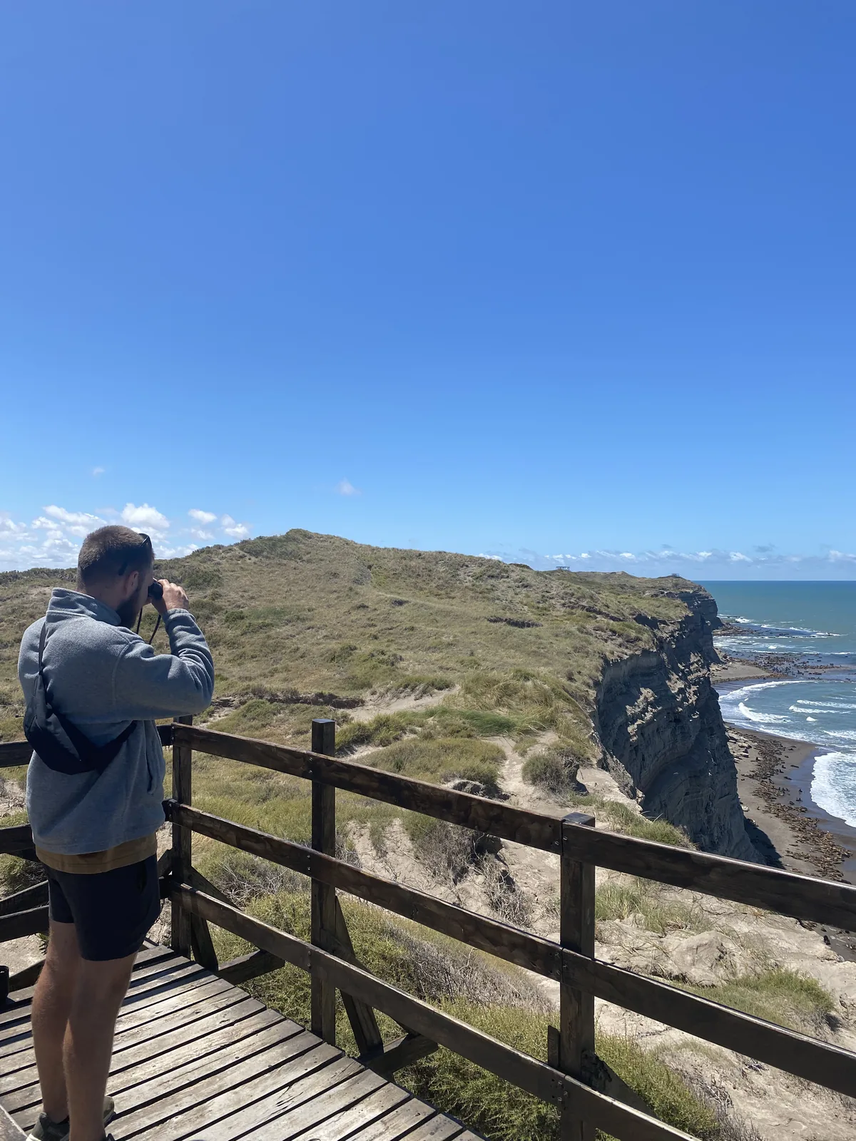 Viajero en mirador de madera con binoculares observando acantilados y el mar en un día soleado de aventura costera
