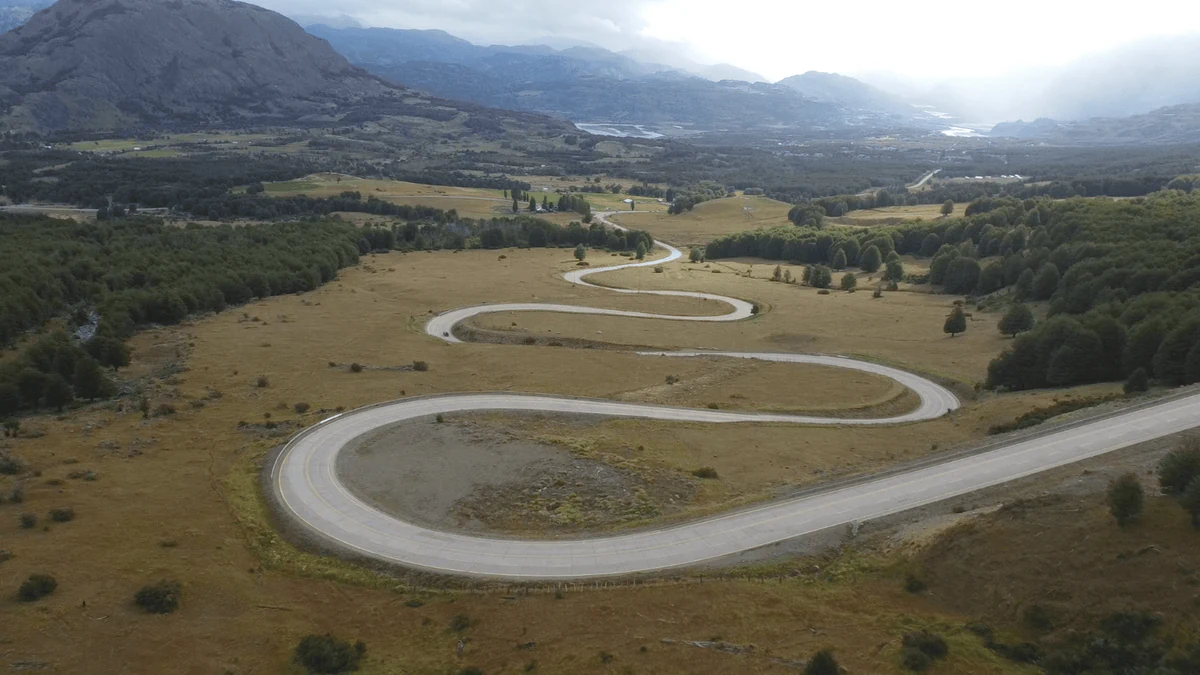 Ruta sinuosa asfaltada serpenteando un valle patagónico, rodeado de campos, bosques y montañas grises