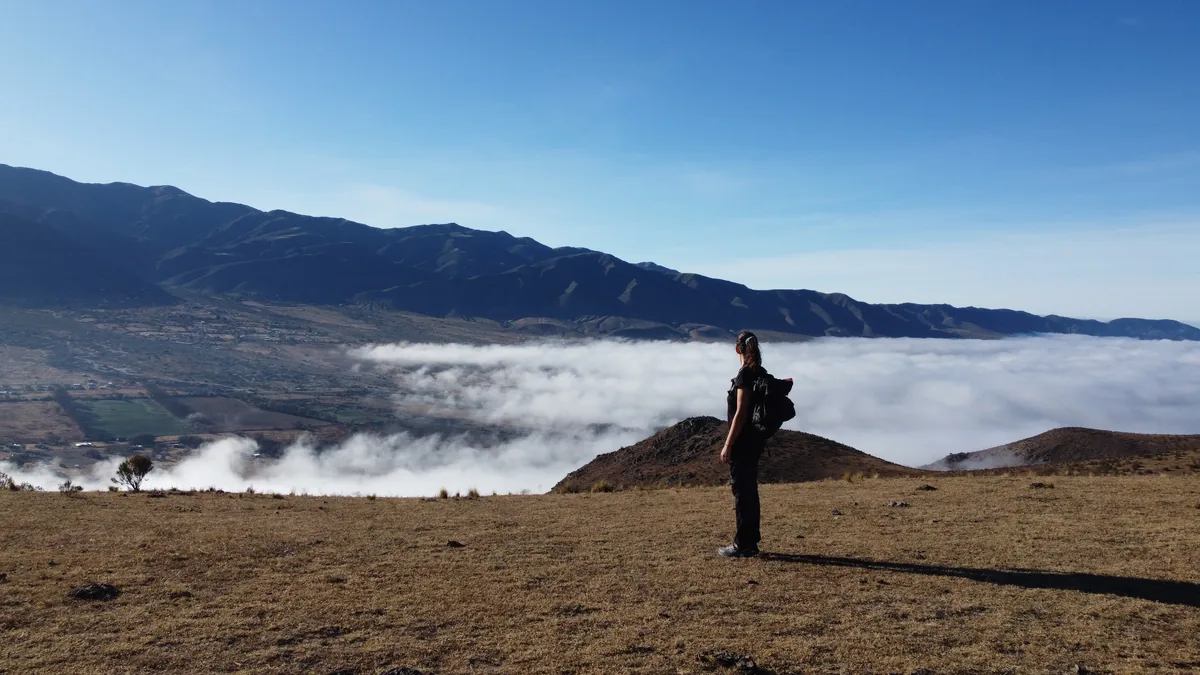 Viajera con mochila en la cima de una colina árida, observando un vasto mar de nubes que cubre un valle montañoso
