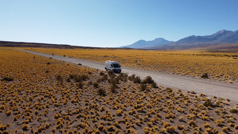Furgoneta camper blanca en camino de ripio, atravesando una vasta estepa árida con pastizales dorados y montañas andinas