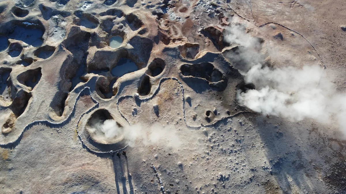 Campo geotérmico con fumarolas de vapor, pozas de agua y un sendero rocoso, con dos vans overland explorando el paisaje